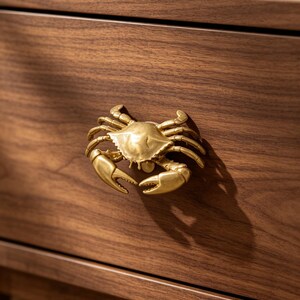 May include: A gold-toned metal cabinet knob shaped like a crab with detailed legs and claws, mounted onto the front of a dark brown wooden drawer. The crab figure is centered on the drawer face, featuring a smooth, metallic finish and texture.