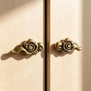 Puede incluir: Dos pomos de armario color latón con forma de nubes estilizadas y patrones en relieve en espiral. Los pomos están montados en una puerta de armario de madera clara con veta vertical, proyectando sombras marcadas a la izquierda.