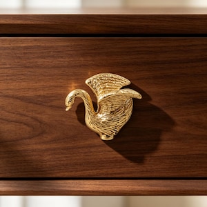 Pode incluir: Um puxador de móvel em metal dourado com formato de cisne com as asas parcialmente abertas, apresentando texturas de penas detalhadas. O puxador de cisne está montado numa frente de gaveta de madeira castanha escura com veio horizontal.