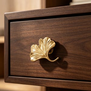 May include: A gold-toned metal drawer pull shaped like a ginkgo leaf attached to a dark brown wooden drawer. The handle features a textured, fan-like surface with distinct veins and a curved stem, providing a functional grip for opening the drawer.