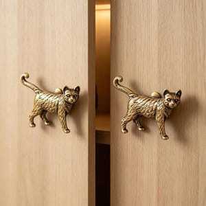 Puede incluir: Dos pomos de armario de metal color latón con forma de gatos de pie y detalles de pelaje texturizado. Cada pomo está montado en una puerta de armario de madera clara, mostrando un gato con la cola arqueada y la cabeza hacia adelante.
