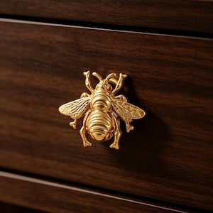 Puede incluir: Un tirador de cajón dorado con forma de abeja, con alas y segmentos corporales detallados, montado en un frente de cajón de madera de color marrón oscuro. El herraje metálico con forma de insecto tiene un acabado pulido y está centrado.