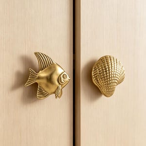 以下が含まれることがあります： 明るい色の木製ドアに取り付けられた、真鍮色の金属製キャビネットノブ2つ。1つはヒレや鱗が細かく表現されたエンゼルフィッシュの形をしており、もう1つは扇状の筋が入った質感のある貝殻の形をしています。