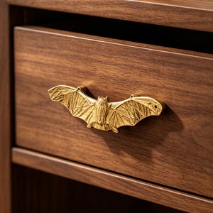 May include: A gold-colored metal drawer pull shaped like a bat with outstretched wings and textured wing details. The bat-shaped handle is mounted horizontally on the front of a dark brown wooden drawer with a visible horizontal wood grain pattern.