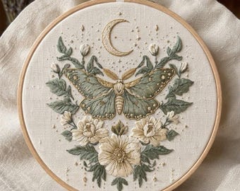 May include: A circular wooden embroidery hoop containing a cream-colored fabric panel. The fabric features detailed hand embroidery of a large sage green moth centered below a crescent moon, surrounded by cream-colored flowers and green leaves.