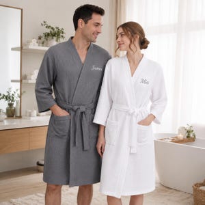 May include: A dark gray waffle-weave robe with the name James embroidered on the chest and a white waffle-weave robe with the name Mia embroidered on the chest. Both robes feature long sleeves, a tie waist, and two front patch pockets.