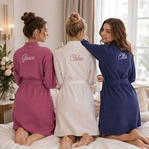 Könnte beinhalten: Drei Waffelpiqué-Bademäntel in Magenta, Weiß und Marineblau. Jeder Bademantel hat einen Bindegürtel, lange Ärmel und einen auf den Rücken gestickten Namen in Schreibschrift: Grace auf dem magentafarbenen, Chloe auf dem weißen und Ella auf dem blauen.