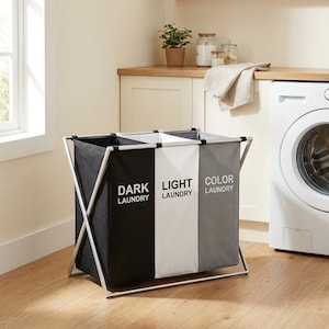 May include: A three-section laundry hamper with a silver metal X-frame. The fabric compartments are black, white, and gray, labeled with the text DARK LAUNDRY, LIGHT LAUNDRY, and COLOR LAUNDRY in white and black lettering respectively.