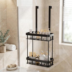 Puede incluir: Un organizador de ducha de metal negro mate con dos estantes rectangulares y barandillas protectoras. La unidad cuelga de dos ganchos verticales diseñados para montarse sobre una puerta de ducha de cristal, conteniendo varios frascos.