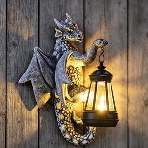 Puede incluir: Una escultura de pared de dragón con textura de piedra gris, escamas y alas detalladas, que sostiene una linterna negra que emite una luz amarilla cálida y brillante. El dragón se muestra en una posición posada con la cola curvada hacia abajo.