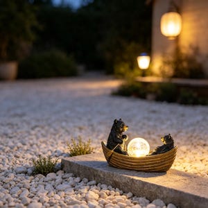 May include: A decorative garden ornament featuring two dark brown bear figurines sitting in a small wooden-textured boat. Between the bears, a glowing, spherical, crackled glass light illuminates the scene with a warm, soft yellow light.