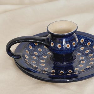 May include: A dark blue ceramic candle holder with a circular base and a side handle. The surface is decorated with a repeating pattern of small white dots with brown centres. The interior of the candle cup is a smooth, light cream-coloured glaze.