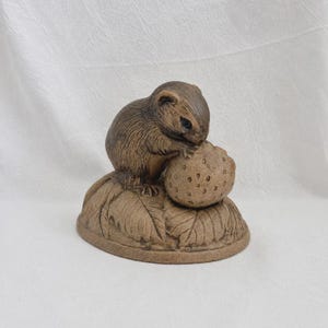 Könnte beinhalten: Eine braune, skulptierte Figur eines kleinen Nagetiers, das auf einer texturierten Blattbasis sitzt. Das Tier hält ein rundes, genopptes Objekt mit beiden Vorderpfoten. Das gesamte Stück hat ein mattes Finish in erdigen Farbtönen.
