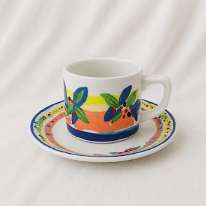 May include: A white ceramic cup and saucer set featuring a hand-painted design with horizontal bands of yellow, orange, and blue. The pattern includes blue leaves and small red and black dots, with a matching decorative border on the saucer rim.