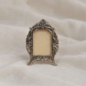 May include: A small, arched picture frame with a metallic, antique silver finish. The frame features intricate, raised floral and leaf detailing along the edges, surrounding a plain, light-coloured rectangular insert in the centre.