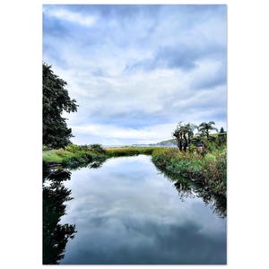 May include: A calm river flows through a landscape with lush green vegetation and trees on both banks. The water surface acts as a mirror, reflecting the cloudy blue sky above. In the distance, the river meets a coastline under a bright horizon.