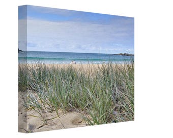 Op de afbeelding: Een rechthoekige canvasprint met een kustlandschap met hoog groen strandgras op de voorgrond, een zandstrand en een kalme blauwe oceaan onder een lichtblauwe lucht met zachte witte wolken. Twee kleine figuren zijn zichtbaar aan de kustlijn.