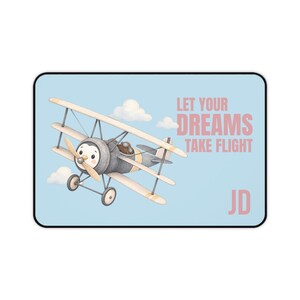 Puede incluir: Una alfombrilla de escritorio rectangular de color azul claro con un biplano de dibujos animados y un pingüino piloto. El texto LET YOUR DREAMS TAKE FLIGHT aparece en rosa, con las iniciales JD en la esquina inferior derecha.