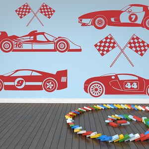 May include: Red race car decals with numbers 7, 9, 16, and 44 on a light blue wall. The decals are arranged in a staggered pattern. There are also two red checkered flags crossed in the upper left corner and two red checkered flags crossed in the upper right corner. The wall is above a wooden floor with a circle of colorful toy blocks.