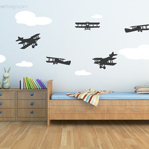 May include: A light blue wall with white clouds and black airplane silhouettes. A wooden bed with a blue mattress and a striped blanket is in the foreground. There are two wooden drawers with blue knobs on either side of the bed.