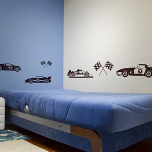 May include: A blue wall with a white wall to the right. A bed with a blue mattress is in the foreground. There are black and white racing car decals on the walls, including a race car with the number 44, a race car with the number 7, and a race car with the number 9. There are also black and white checkered flags on the walls.