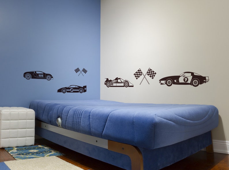 May include: A blue wall with a white wall to the right. A bed with a blue mattress is in the foreground. There are black and white racing car decals on the walls, including a race car with the number 44, a race car with the number 7, and a race car with the number 9. There are also black and white checkered flags on the walls.
