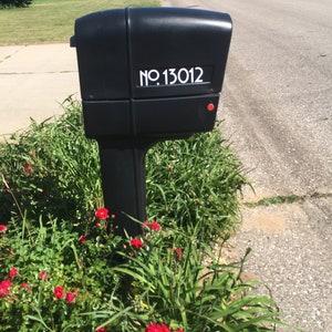 May include: A black mailbox with a white number sticker that reads "NO. 13012". The mailbox is mounted on a post and surrounded by green grass and red flowers.