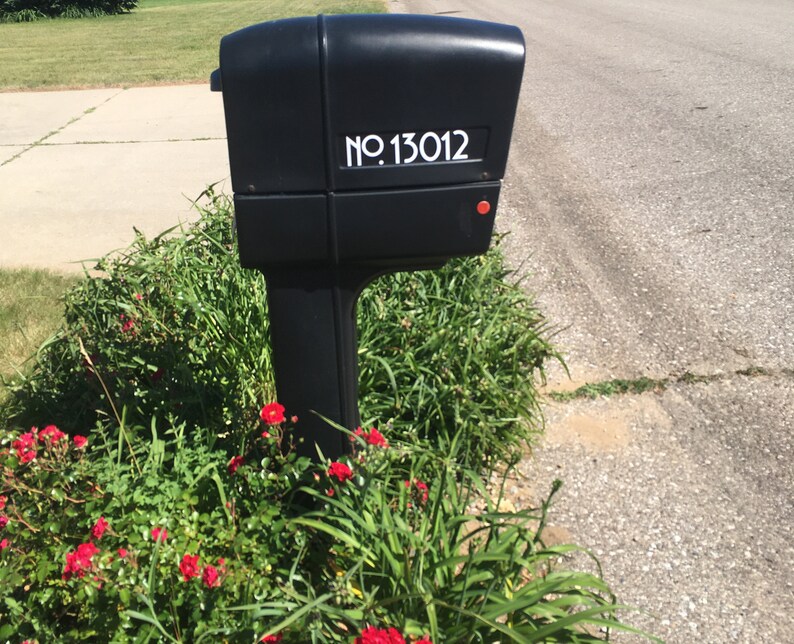 May include: A black mailbox with a white number sticker that reads "NO. 13012". The mailbox is mounted on a post and surrounded by green grass and red flowers.