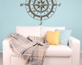 May include: A white couch with a brown and white plaid throw blanket and yellow and blue striped pillows. A brown compass with the words "N", "S", "E", "W", "NW", "NE", "SW", "SE" is on the wall behind the couch.