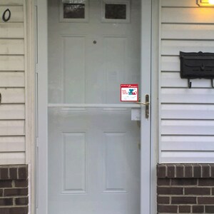 May include: White front door with a glass storm door and a small red sticker that says "In Case of Emergency, Please Call Our Vet's" with a phone number. The door has a brass door handle and a brass deadbolt lock.