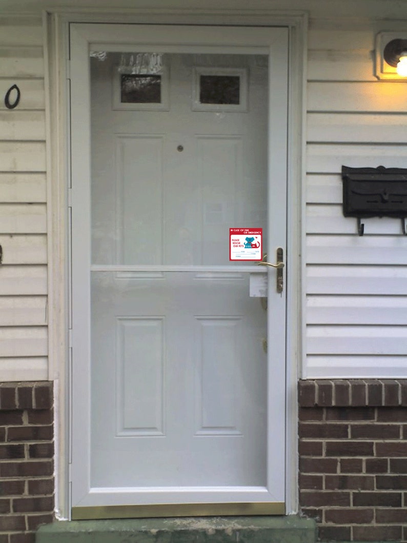 May include: White front door with a glass storm door and a small red sticker that says "In Case of Emergency, Please Call Our Vet's" with a phone number. The door has a brass door handle and a brass deadbolt lock.