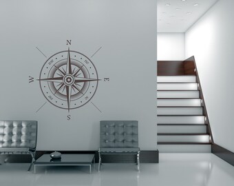 May include: A brown compass with the directions north, south, east, and west, and the cardinal points NW, NE, SW, and SE. The compass is on a white wall in a modern living room with two gray chairs and a black coffee table.