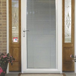 May include: White front door with a single pane of glass and a white door frame. The glass has white blinds inside. The door has a silver door handle and a small red and blue sticker on the door frame.