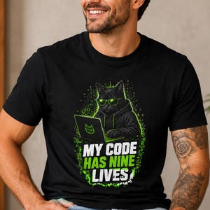 Op de afbeelding: Een zwart t-shirt met korte mouwen en een afbeelding van een zwarte kat met een groene bril en een hoodie achter een laptop. De tekst MY CODE HAS NINE LIVES staat in het wit en groen onder de kat, omringd door groene digitale accenten.