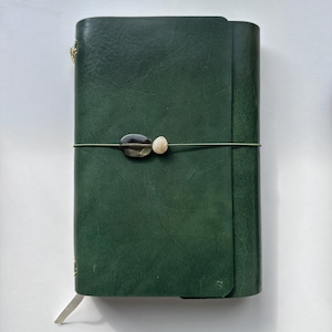 Könnte beinhalten: Ein dunkelgrünes Notizbuch aus Leder mit einem umlaufenden Verschluss, der durch eine dünne elastische Kordel gesichert ist. Die Kordel ist mit zwei glatten, ovalen Perlen verziert, einer dunkelgrau durchscheinenden und einer cremefarbenen.