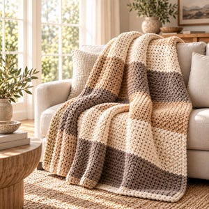 May include: A chunky knit throw blanket featuring a horizontal striped pattern in alternating shades of cream, beige, and dark brown. The blanket has a thick, textured stitch and is draped over the seat and back of a light-colored sofa.