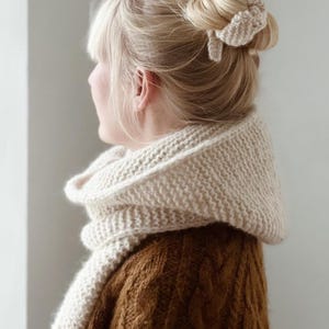 May include: A cream-coloured chunky knit scarf draped around the neck, paired with a matching cream-coloured knit hair scrunchie worn in a bun. The scarf features a thick, textured garter stitch pattern, and the hair accessory has a similar knit texture.