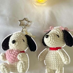 May include: Two handmade crochet dog figures with white bodies, black ears, and red collars. One figure holds a pink heart and features a silver star-shaped keychain attachment, while the other figure has small pink bows on its head.