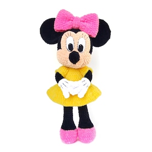 Puede incluir: Una muñeca de peluche tejida a ganchillo que representa un personaje con orejas negras, un lazo rosa brillante y una cara color melocotón. La muñeca lleva un vestido amarillo, guantes blancos y zapatos rosas. La textura es suave y de lana, con ojos negros y blancos.