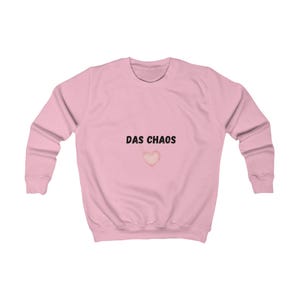 Può includere: Una felpa a maniche lunghe rosa chiaro con il testo nero "DAS CHAOS" stampato al centro del petto, posizionato direttamente sopra una piccola grafica a forma di cuore dai toni rosa tenui. Il capo presenta un girocollo e polsini a coste.