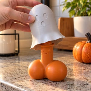 Puede incluir: Una cubierta gris claro con forma de fantasma colocada sobre una figura naranja de forma fálica con una pequeña cara sonriente. La cubierta de fantasma presenta dos ojos ovalados y una boca circular, mientras que la base naranja tiene dos lóbulos redondeados.