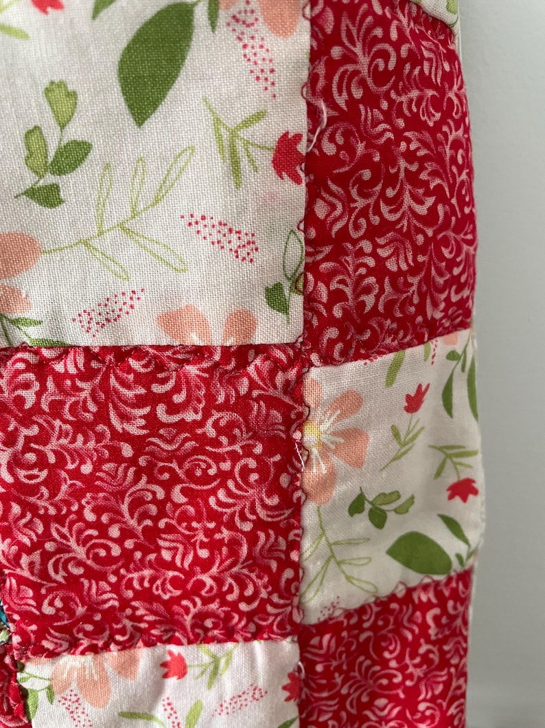 Pu&ograve; includere: Un tessuto patchwork caratterizzato da una griglia ripetitiva di quadrati di stoffa alternati. Un set di quadrati mostra uno sfondo bianco con un delicato motivo floreale nei toni del verde, rosa e rosso. L'altro set presenta uno sfondo rosso tinta unita con una stampa bianca di viticci e foglie vorticose.