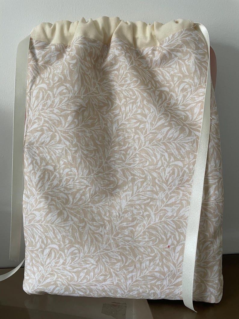 Pu&ograve; includere: Una borsa rettangolare con coulisse in tessuto beige chiaro con un motivo ripetuto di foglie e viticci bianchi. La parte superiore &egrave; in tessuto color crema tinta unita con un'apertura arricciata, chiusa da due lunghi nastri di raso bianco.