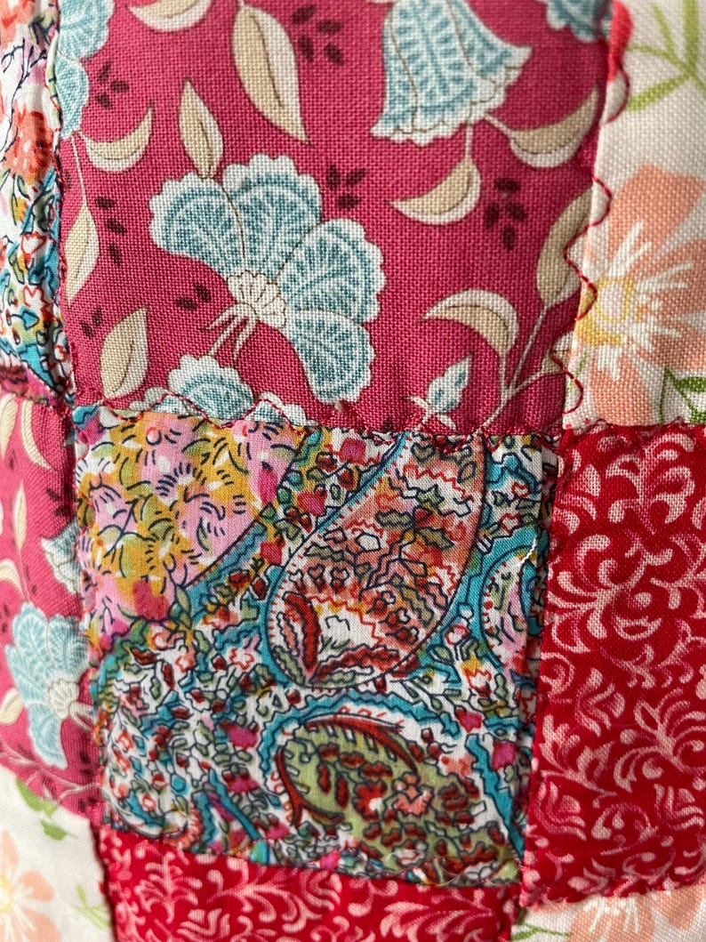 Pu&ograve; includere: Un tessuto patchwork caratterizzato da una variet&agrave; di motivi floreali e paisley in vivaci tonalit&agrave; di magenta, turchese, rosso e crema. I quadrati di tessuto sono uniti da cuciture visibili, creando una superficie strutturata e multicolore.