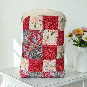 Pu&ograve; includere: Una borsa con coulisse realizzata in tessuto patchwork con una griglia di quadrati rossi, rosa e bianchi con motivi floreali, paisley e a vortice. La borsa ha una parte superiore color crema con nastri bianchi e un'etichetta Hand Made.