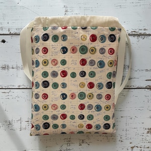 May include: A drawstring bag featuring a cream-coloured fabric printed with a grid pattern of handwritten notes, numbers, and various colourful buttons in shades of red, blue, green, yellow, and grey, finished with plain cream-coloured fabric straps.