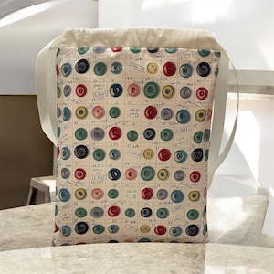 May include: A tote bag featuring a pattern of colourful buttons on a cream background. The buttons are in shades of red, green, blue, and yellow. The bag has cream straps and a cream lining. The bag's dimensions are approximately 30 cm x 25 cm.