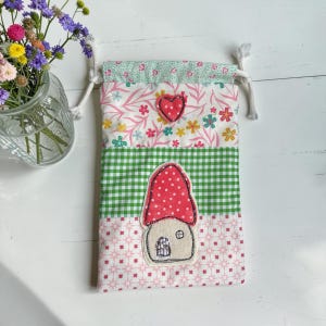 May include: A fabric drawstring pouch featuring a patchwork design with a green gingham section, a floral print, and a pink star pattern. An appliqued red mushroom house with white polka dots and a small red heart are stitched onto the front.