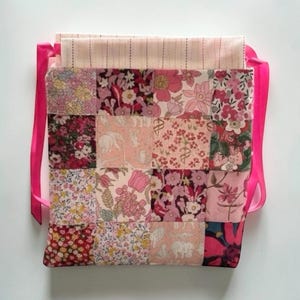 May include: A patchwork tote bag featuring a variety of floral patterns in pink, red, and cream. The bag has a rectangular shape with a pink ribbon handle. The bag is made of fabric and is likely handmade.