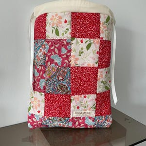 Pu&ograve; includere: Una borsa rettangolare con coulisse caratterizzata da un design patchwork con motivi floreali e paisley rossi, rosa e bianchi. La parte superiore presenta un bordo in tessuto color crema con nastri bianchi. Un'etichetta bianca riporta Hand Made with love.